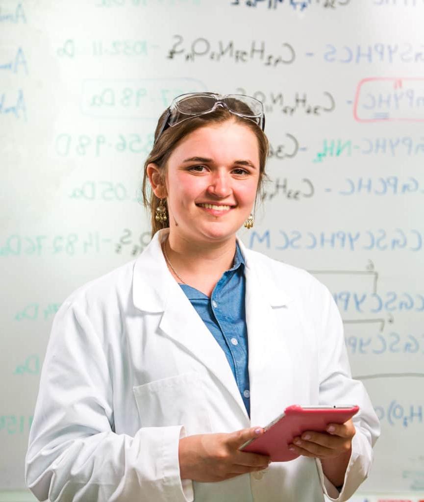 一个穿着实验服的学生站在写满化学代码的白板前. 